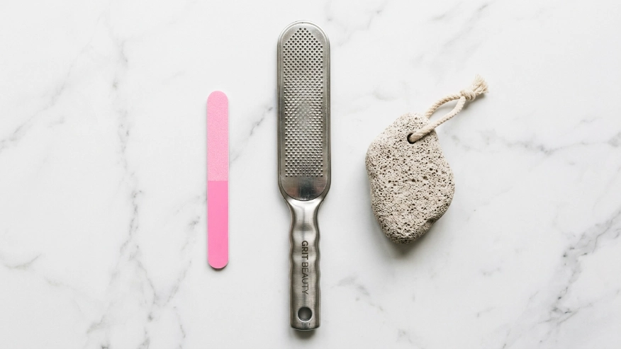 A flat lay photo on a clean white marble background showing a small pink fingernail file next to a large stainless steel foot rasp and a natural pumice stone, bright soft lighting, high-resolution beauty blog photography.

Recommended Size: 900x600 px